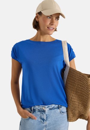 Kobieta w beżowej czapce i niebieskim t-shircie, jasnoniebieskich dżinsach, niesie na ramieniu plecioną brązową torbę typu tote.