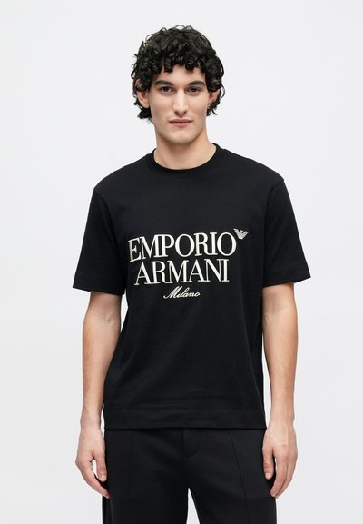 Μαύρο βαμβακερό t-shirt με τη φράση "EMPORIO ARMANI Milano" σε μεγάλο εκρού κείμενο και μικρό λογότυπο στο στήθος. Κοντά μανίκια και στρογγυλή λαιμόκοψη.