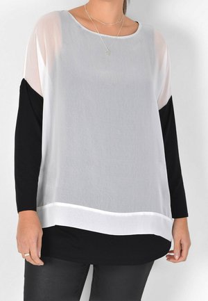 Nainen, joka pukeutunut löysään valkoiseen ja mustaan kerrospaitaan, jossa on läpikuultavat valkoiset ylähihoit ja mustat istuvaiset housut.