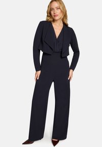 Femme portant une combinaison noire à manches longues avec jambes larges et une veste courte drapée, debout devant un fond blanc uni.