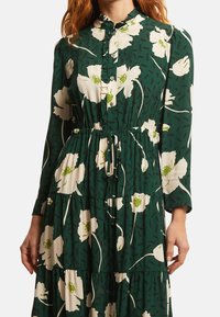 Grön blommig klänning med långa ärmar, knäppning fram och elastisk midja. Har vita blommor och gröna detaljer på ett texturerat tyg.