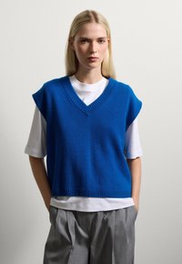 Blauwe gebreide vest met V-hals, mouwloos ontwerp en geribbelde zoom, gedragen over een wit T-shirt en gecombineerd met lichtgrijze geplooide broek.
