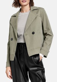 Krótka kurtka w jasnym oliwkowym kolorze z kołnierzem koszulowym, zapinana na dwa rzędy czarnych guzików. Noszona na paskowanej koszuli i czarnych spodniach.