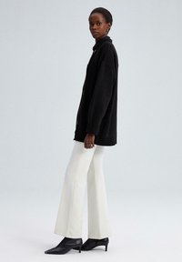Vrouw in een zwart oversized knopen cardigan, witte flare broek en zwarte puntige haklaarzen, staand tegen een effen achtergrond.