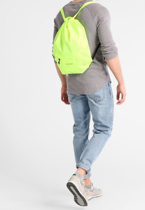 Personne marchant à l'écart portant une chemise à manches longues gris clair, un jean bleu clair, des baskets beiges et un sac à dos à cordon vert néon.