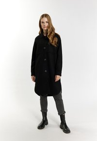 Černé oversized šaty košového střihu s límečkem, předním zapínáním na knoflíky a dvěma náprsními kapsami. Mají dlouhé rukávy a zaoblený spodní lem. V kombinaci s černými kotníkovými botami.