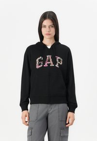 Black zip-up hoodie featuring floral "GAP" lettering, adjustable drawstring hood, and front pockets. Soft fabric with ribbed cuffs.