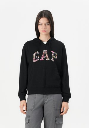Svart zip-up hettegenser med blomstrete "GAP" bokstaver, justerbar snor i hetten og frontlommer. Mykt stoff med ribbede mansjetter.