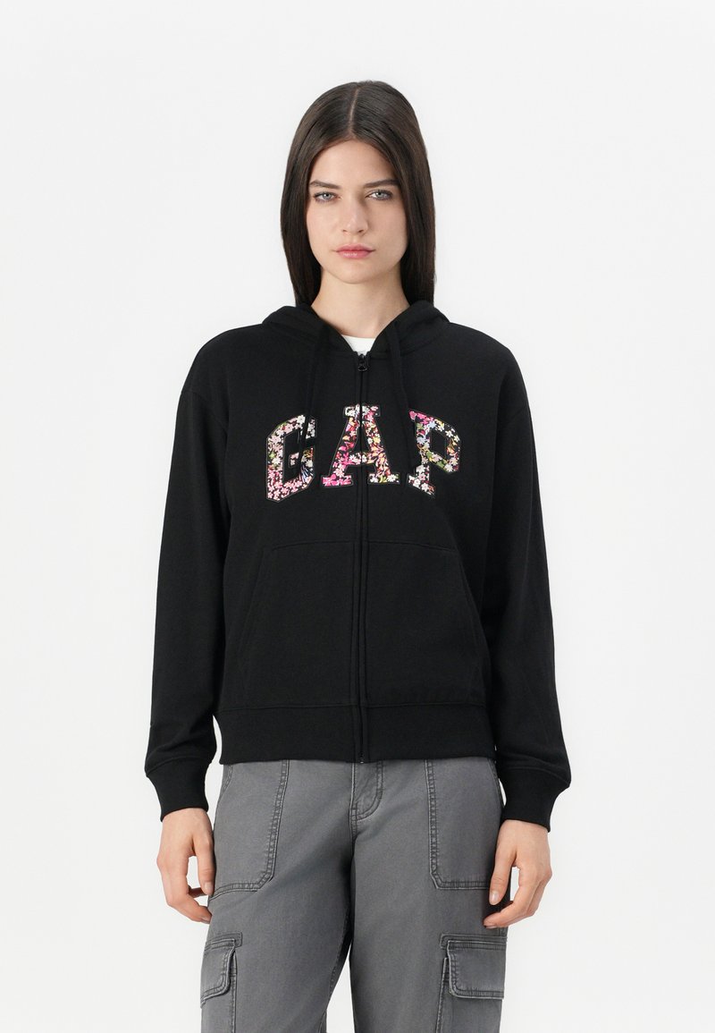 Svart zip-up hettegenser med blomstrete "GAP" bokstaver, justerbar snor i hetten og frontlommer. Mykt stoff med ribbede mansjetter.