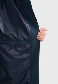 Veste bleu marine avec une doublure intérieure lisse et brillante et une poche latérale, tenue par une main. Le tissu semble léger et légèrement froissé.