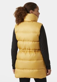 Žlutá zateplená vesta s vysokým límcem, s nařaseným pasem, prošívanou texturou a černými dlouhými rukávy pod ní.