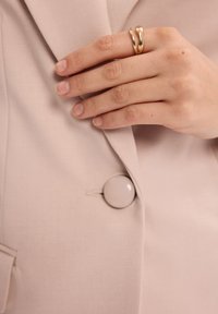 Jasnoróżowa marynarka z gładkiego materiału, z dużym okrągłym guzikiem i ręką noszącą złoty podwójny pierścionek umieszczoną na klapie.