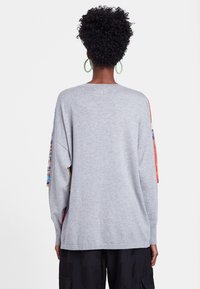 Grå oversized tröja med långa ärmar som har färgglada mönstrade detaljer på axlarna. Mjuk textur, ribbade ärmslut och nederkant.