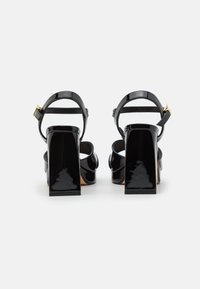 Zwarte lakleren sandalen met hoge hakken, een chunky vierkante hak, enkelriem en een gouden gespdetail.