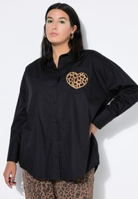 Black button-up shirt with a leopard-print heart patch on the chest. Loose fit, long sleeves, and smooth fabric. Patterned pants visible below.