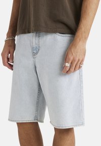 Shorts de mezclilla azul claro con un ajuste holgado, que presentan piernas rectas, costuras visibles y bolsillos laterales. Combinados con una camiseta marrón.