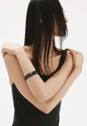 Mujer con camiseta sin mangas negra cruzando los brazos, lleva una pulsera negra; cabello largo y lacio que cubre parcialmente su rostro, fondo blanco.