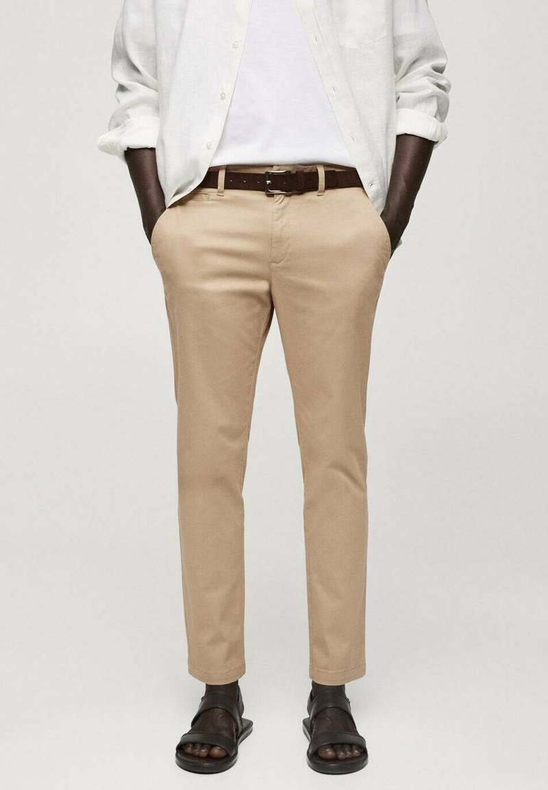 Homme portant un pantalon slim beige, une chemise blanche avec les manches retroussées, une ceinture marron et des sandales noires, les mains dans les poches, debout devant un fond uni.