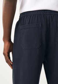 Tamsiai mėlynos kelnės iš minkštos medžiagos, su elastinga juosmens juosta, šoninėmis siūlėmis kišenėmis ir viena kvadrato formos kišene gale.