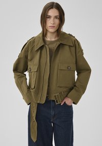 Frau trägt eine olivgrüne, verkürzte Jacke mit großen, geknöpften Taschen und Gürtel, kombiniert mit dunkelblauen Jeans, steht vor einem grauen Hintergrund.