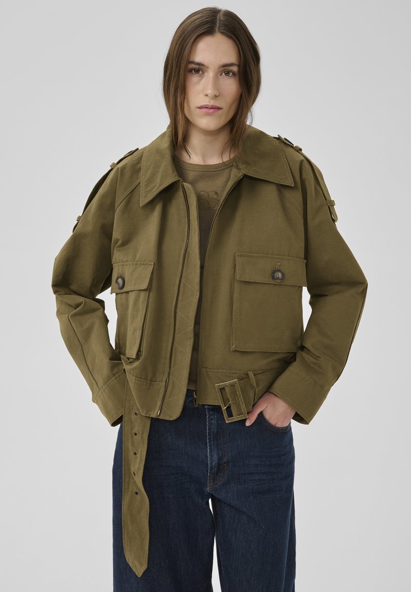 Frau trägt eine olivgrüne, verkürzte Jacke mit großen, geknöpften Taschen und Gürtel, kombiniert mit dunkelblauen Jeans, steht vor einem grauen Hintergrund.