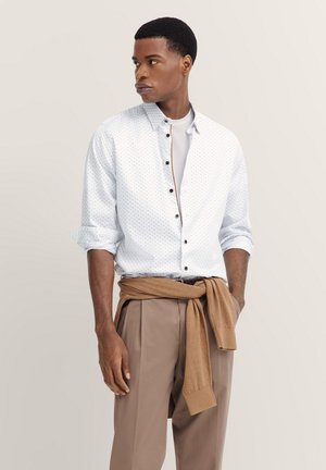 Weißes Hemd mit blauen Punkten, hochgekrempelte Ärmel und ein brauner Pullover um die Taille gebunden. Beigefarbene Hosen runden das Outfit ab.