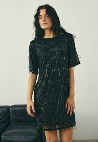 Schwarzes Pailletten-Kleid mit lockerem Sitz, kurzen Ärmeln und einem strukturierten Fransen-Design entlang des Saums, das einen schimmernden Effekt erzeugt.