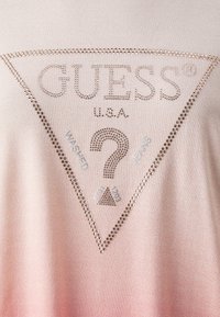 T-shirt con gradiente rosa e accenti di strass. Presenta un logo triangolare con "GUESS U.S.A." e il testo "WASHED JEANS" in argento.