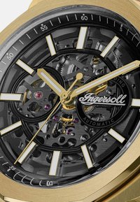 Primo piano di un orologio automatico dorato con ingranaggi visibili, marcatori delle ore bianchi e il logo Ingersoll su un quadrante scheletrato nero.