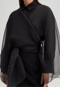 Sort gennemsigtig omslag med en blød tekstur, der har brede ærmer og et taljebånd, over en matchende top. Simpelt, elegant design med minimal dekoration.