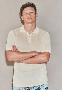 Polo jaune pâle à manches courtes avec une texture en maille et une patte de boutonnage à trois boutons. Le modèle pose les bras croisés, portant un short à motifs.