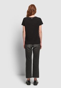 Mujer con cabello largo y rojo, vestida con una camiseta negra de manga corta, jeans gris oscuro y zapatos negros, de pie de espaldas sobre un fondo liso.
