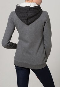 Persona vestida con una sudadera gris con una capucha de punto negra forrada con felpa blanca, combinada con pantalones vaqueros ajustados de color azul oscuro, vista desde atrás.