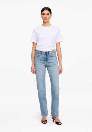 Femme aux cheveux foncés portant un t-shirt blanc, un jean droit taille haute bleu clair et des talons ouverts noirs, debout devant un fond blanc.