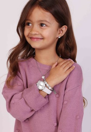 Zilveren horloge met een witte band, zilveren oorbellen in de vorm van hartjes en een bijpassende ketting; meisje draagt een roze textuurtrui.