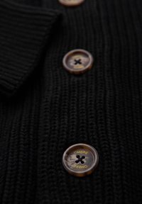 Czarny sweter z dzianiny z teksturowanej tkaniny, dużymi bursztynowymi guzikami z czarnym przeszyciem oraz napisem "OMBRE" na każdym guziku.