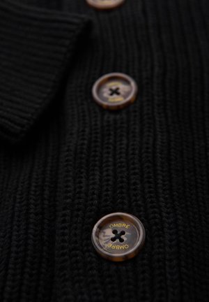 Schwarze Strickjacke aus strukturiertem Stoff mit großen bernsteinfarbenen Knöpfen und schwarzer Naht sowie dem Aufdruck "OMBRE" auf jedem Knopf.