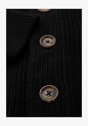 Schwarze Strickjacke aus strukturiertem Stoff mit großen bernsteinfarbenen Knöpfen und schwarzer Naht sowie dem Aufdruck "OMBRE" auf jedem Knopf.