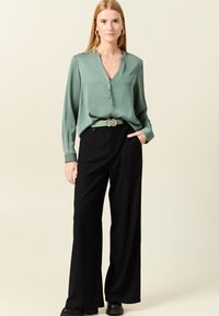 Groene zijden blouse met een knoopsluiting, gecombineerd met zwarte wijde broek met hoge taille en een lichtgroene riem. Zwart schoeisel accentueert de outfit.
