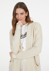 Beige Strickjacke mit offenem Vorderteil, langen Ärmeln, gerippten Bündchen und Seitentaschen, getragen über einem weißen T-Shirt mit schwarzer Schrift.