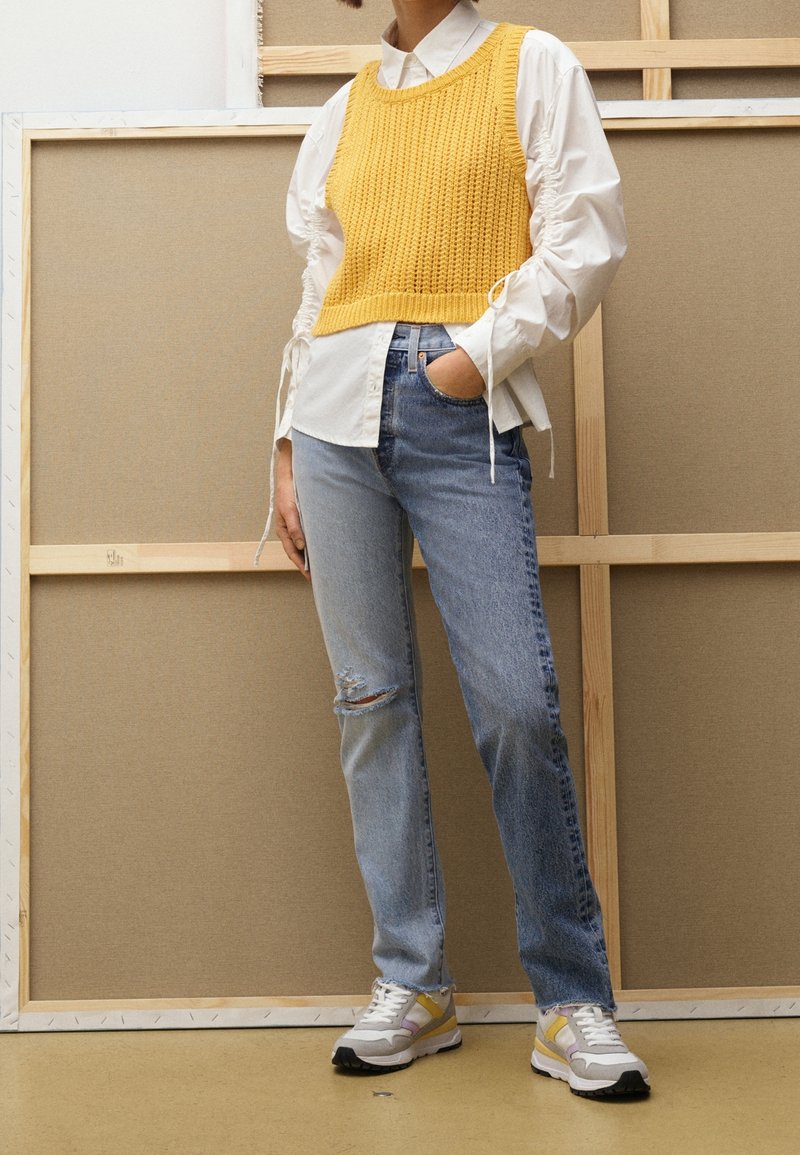 Gul stickad linne över en vit skjorta, parat med ljusblå slitna jeans och grå sneakers med gula accenter.