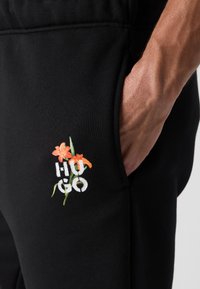 Pantalons de jogging en coton noir avec poches latérales, arborant le texte brodé "HUGO" et des accents floraux colorés sur la zone de la poche gauche.