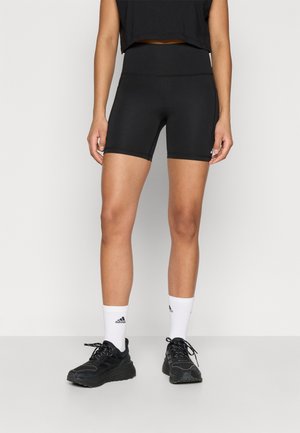 Kvinde iført sorte højtaljede biker-shorts, hvide Adidas-strømper og sorte atletiksk sko, stående mod en ensfarvet baggrund.