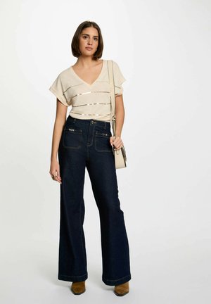 Top beige à col en V avec des rayures horizontales, associé à un jean large bleu foncé. La mannequin tient un petit sac bandoulière crème, portant des bottines à talons marron.