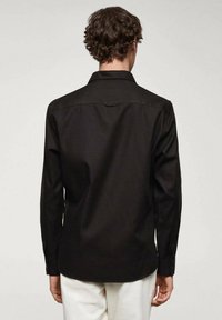 Czarna koszula z długim rękawem z klasycznym kołnierzykiem i detalem na plecach, wykonana z gładkiego materiału. Koszula jest wsunięta w jasne spodnie.