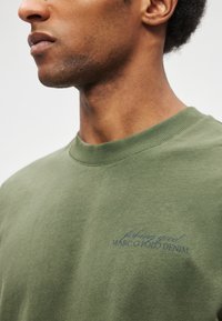 Sweatshirt verde-oliva feita de algodão, com gola redonda e punhos canelados. A frente exibe um texto bordado azul: "fishing good MARC O POLO DENIM."