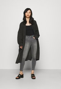 Cardigan preto, de malha larga, por cima de um top preto, combinado com jeans skinny cinzas e sandálias pretas de salto com detalhe de fivela.