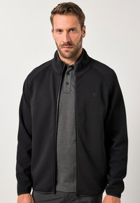 Giacca nera con zip e colletto alto, caratterizzata da una texture liscia e un logo discreto. Indossata sopra una polo grigia scura con bottoni.