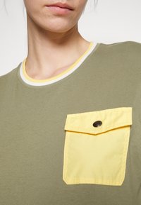 Olivově zelené tričko s žlutou klopovou kapsou. Má žebrovaný výstřih s žlutými a bílými pruhy. Měkčená textilie.