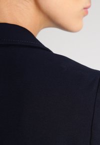 Blazer aderente blu navy in tessuto liscio, con dettagli di cucitura sottili e colletto classico, mostrato di profilo e da dietro.